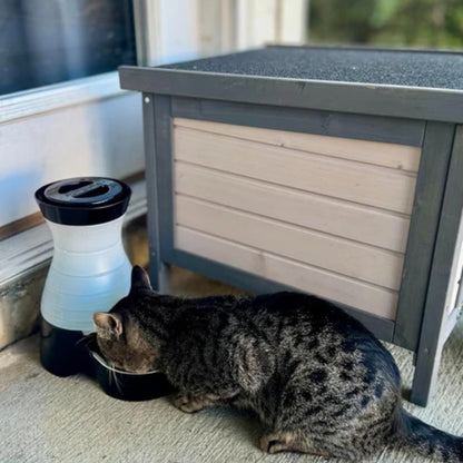 A dark tabby cat drinks from a black and white pet waterer next to a gray and beige storage box on a porch.